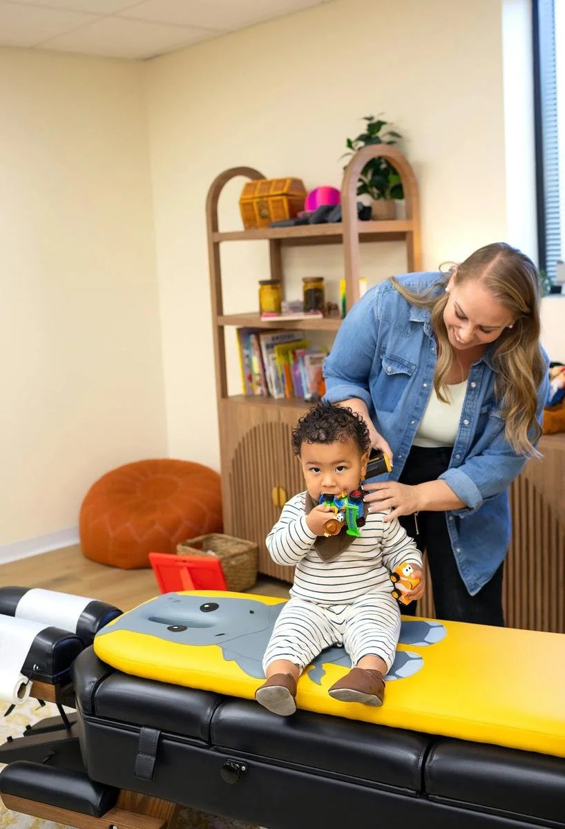 Toddler sitting with toys on a pediatric chiropractic table while a chiropractor stands nearby